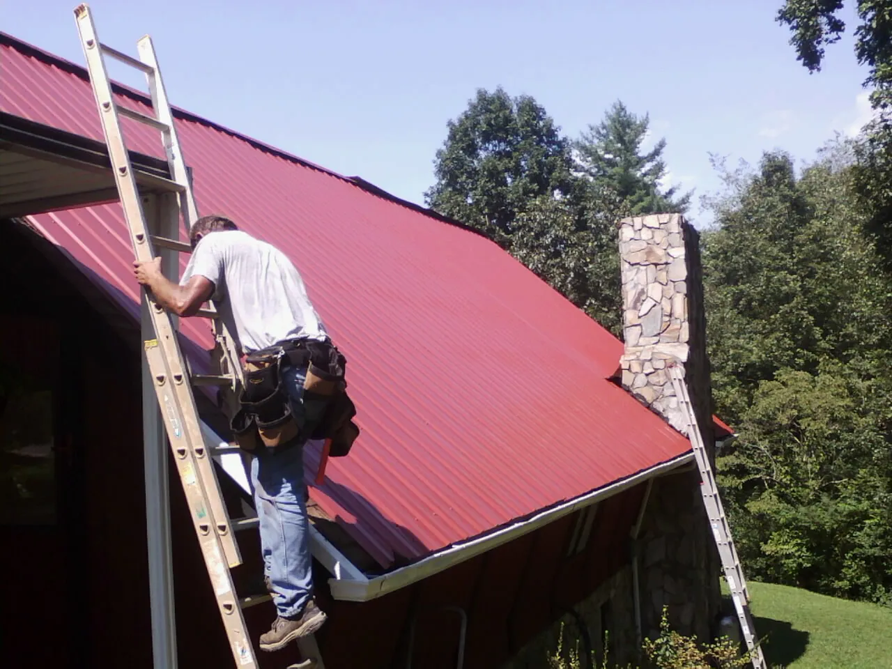 Roofer on a ladder beside a red metal roof and stone chimney — Soffit & Fascia Repair by Roofing Brothers in New Bloomington